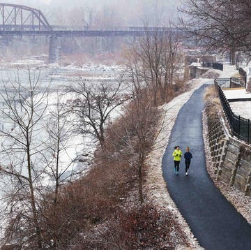 Two individuals jogging along a snowcovered path beside a river with a bridge in the background two individuals jogging along a snowcovered path beside a river with a bridge in the background