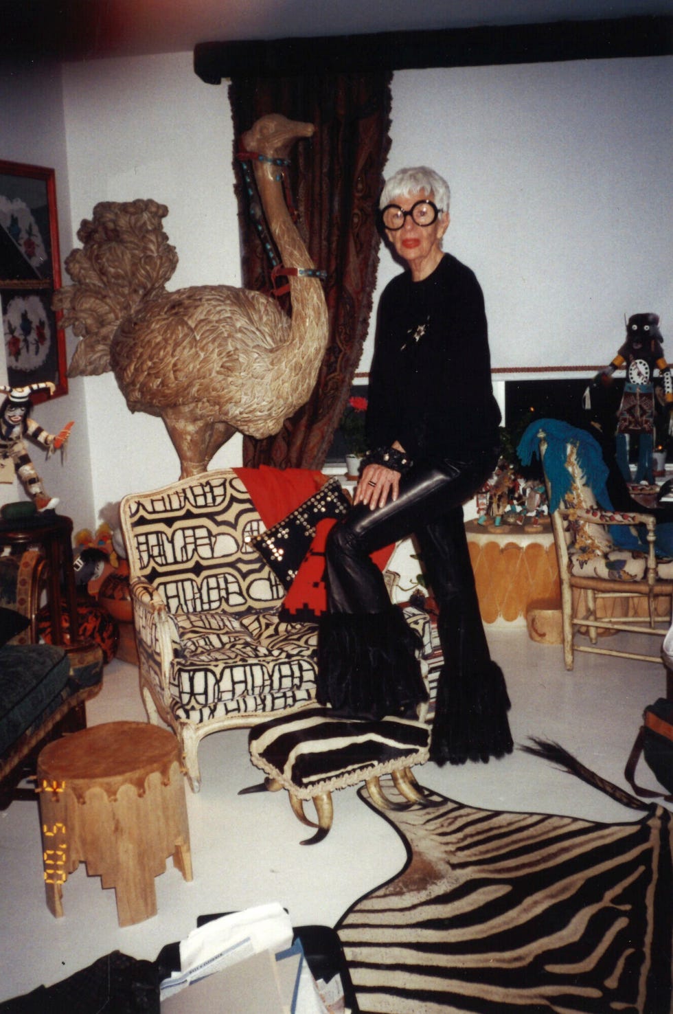 A person in distinctive attire is seated on a patterned couch with a large decorative ostrich statue beside them