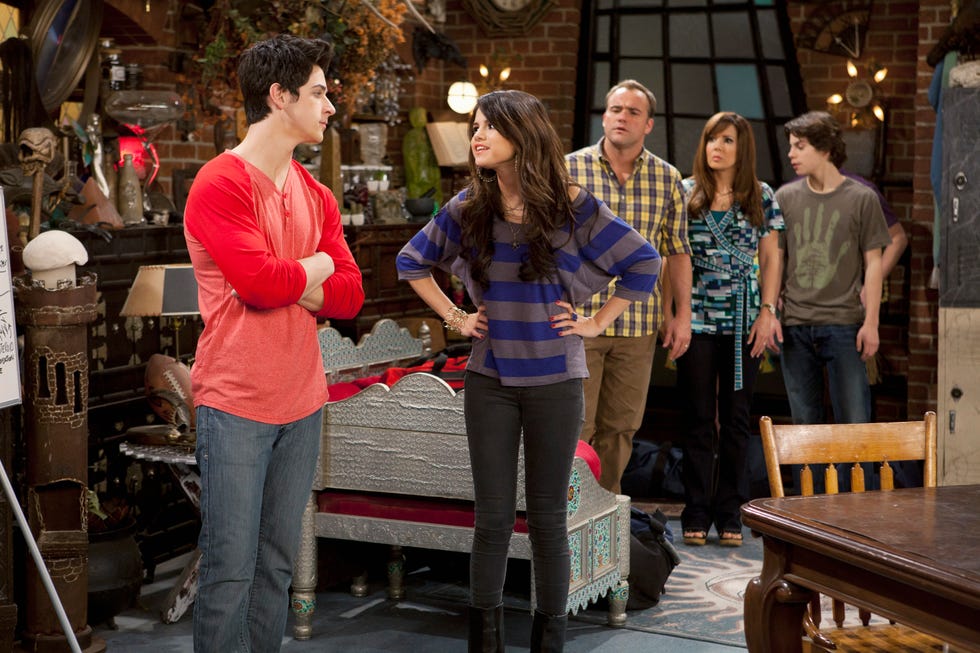 selena gomez stands with her hands on her hips in character for wizards of waverly place, she looks at a costar as other actors stand in the background