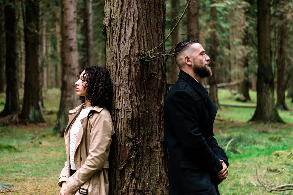 a woman and a man standing leaning each side against a tree in the forest
