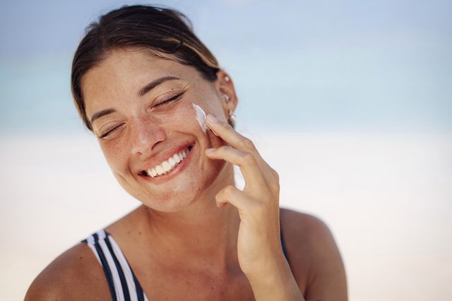 una mujer con un banador de rayas se aplica crema en la cara mientras sonrie
