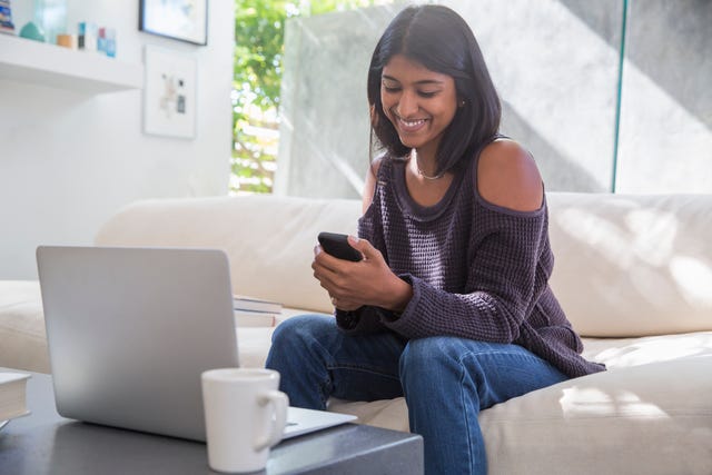 Woman at laptop texting with cell phone on living room sofa