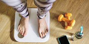 woman checking weight while standing on weight scale by dumbbell, water bottle and mobile phone at home