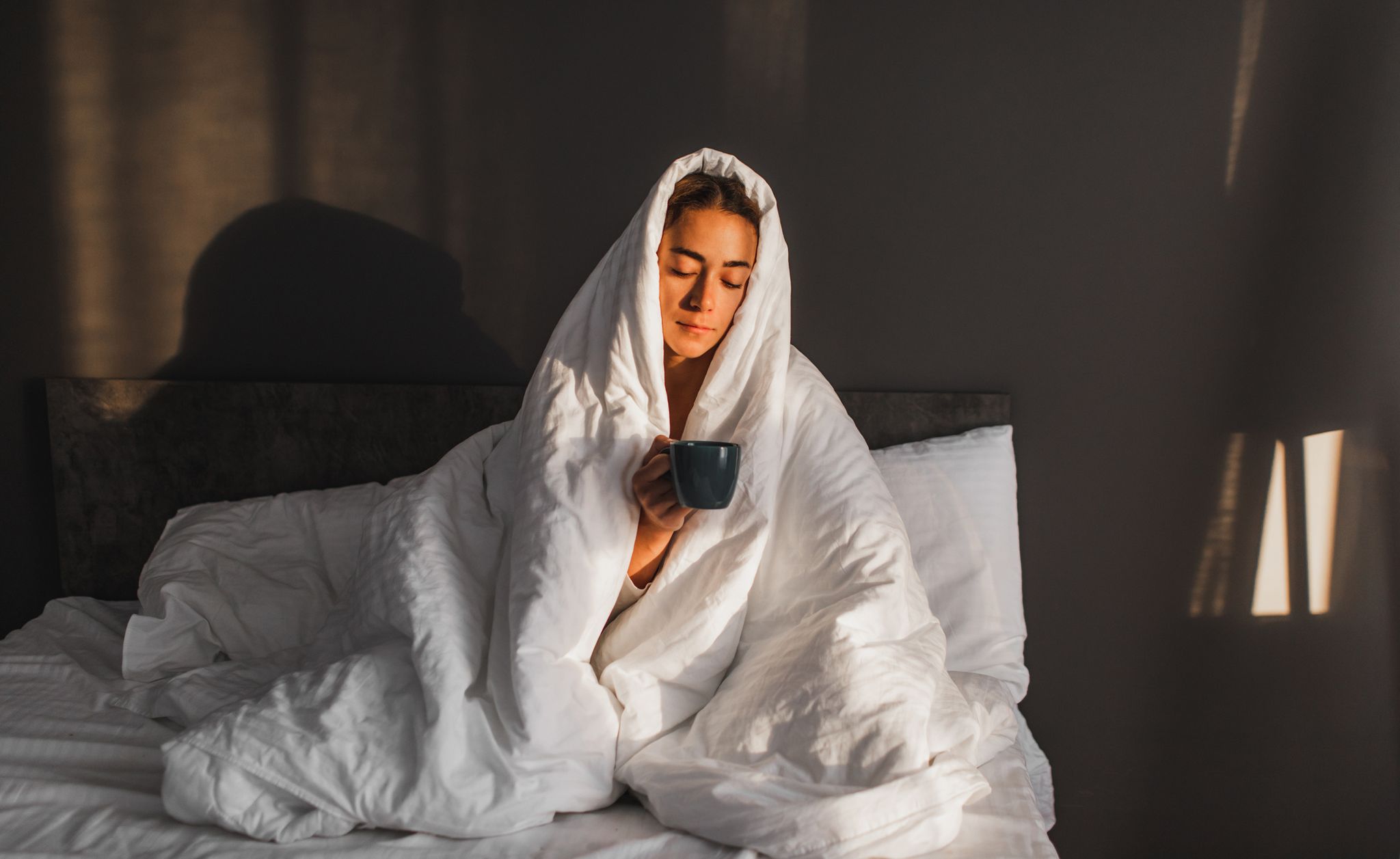 woman wrapped in blanket in bed holding a hot drink
