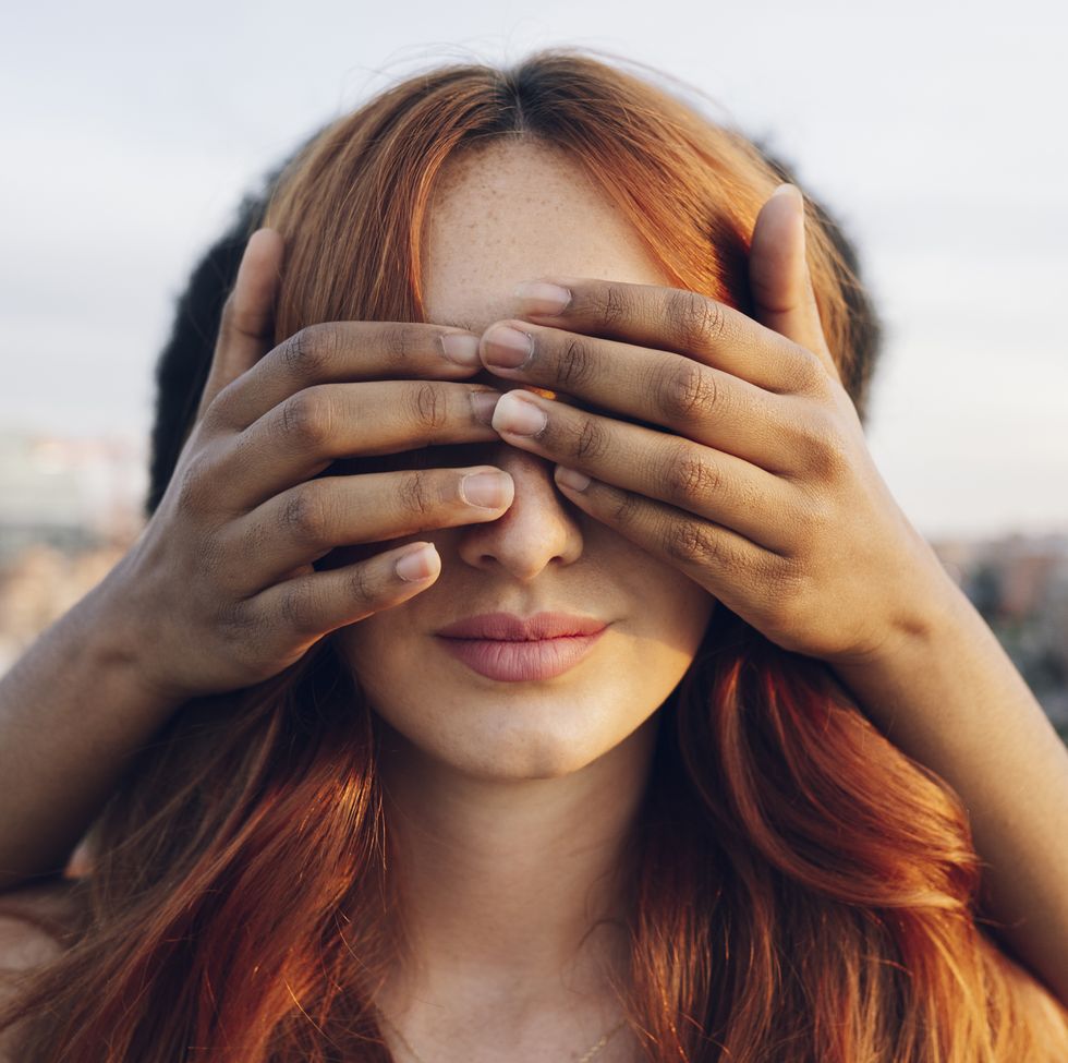 woman covering eyes of redhead girlfriend with hands at sunset