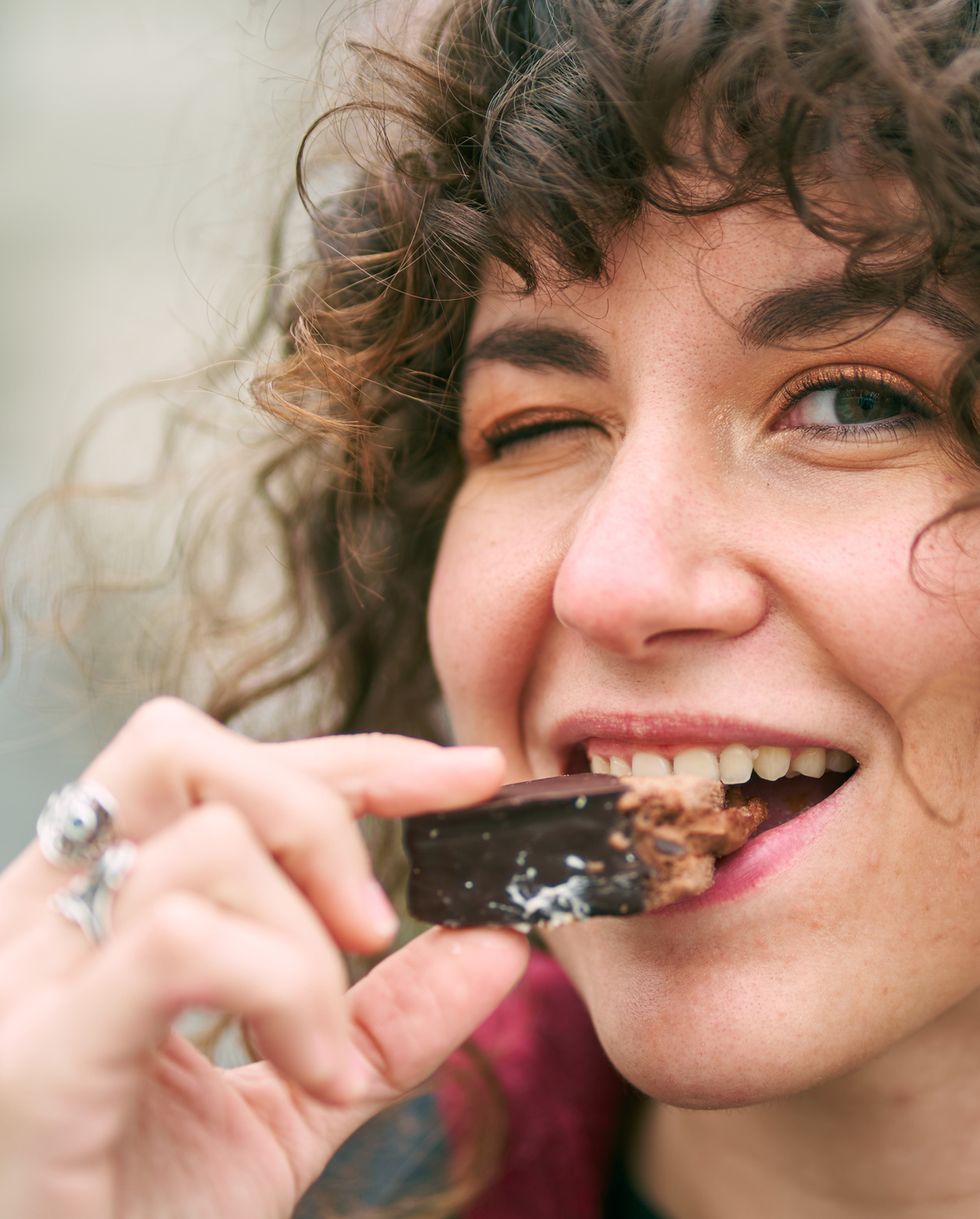 positive female with curly hair in headband winking and looking at camera while eating sweet chocolate candy against blurred background