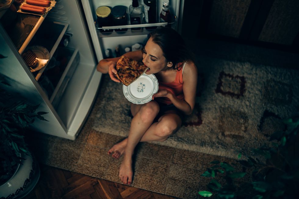 Woman eating pizza slice in front of the refrigerator