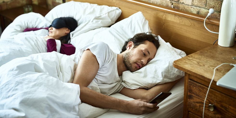 woman fast asleep next to partner who is checking his smartphone