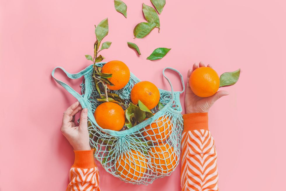 woman hands in orange striped sweatshirt holding oranges with green leaves in blue eco friendly shopping mesh bag on pink background