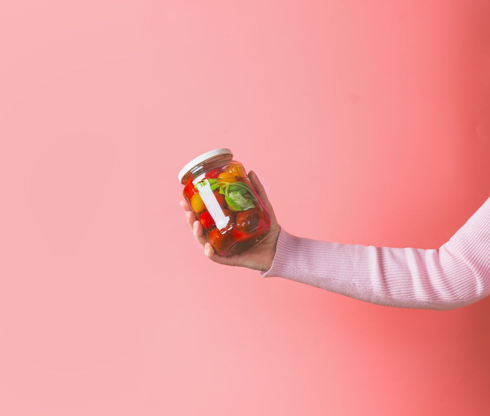 woman hands with light pink sweatshirt holding glass jar with various preserved food on pink background