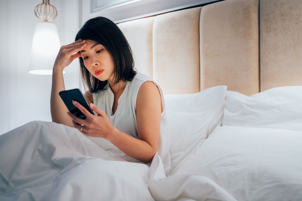 woman holding mobile phone feeling headache sitting on bed at home