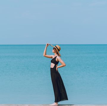 a woman in a black bikini, wearing a long black skirt and wearing a hat, stands by the sea and the sky is blue