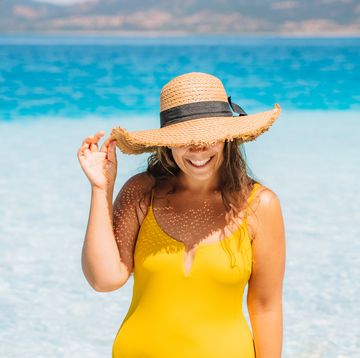 a woman in a yellow swimsuit and straw hat emerges from the sea with clear turquoise water on a summer sunny day