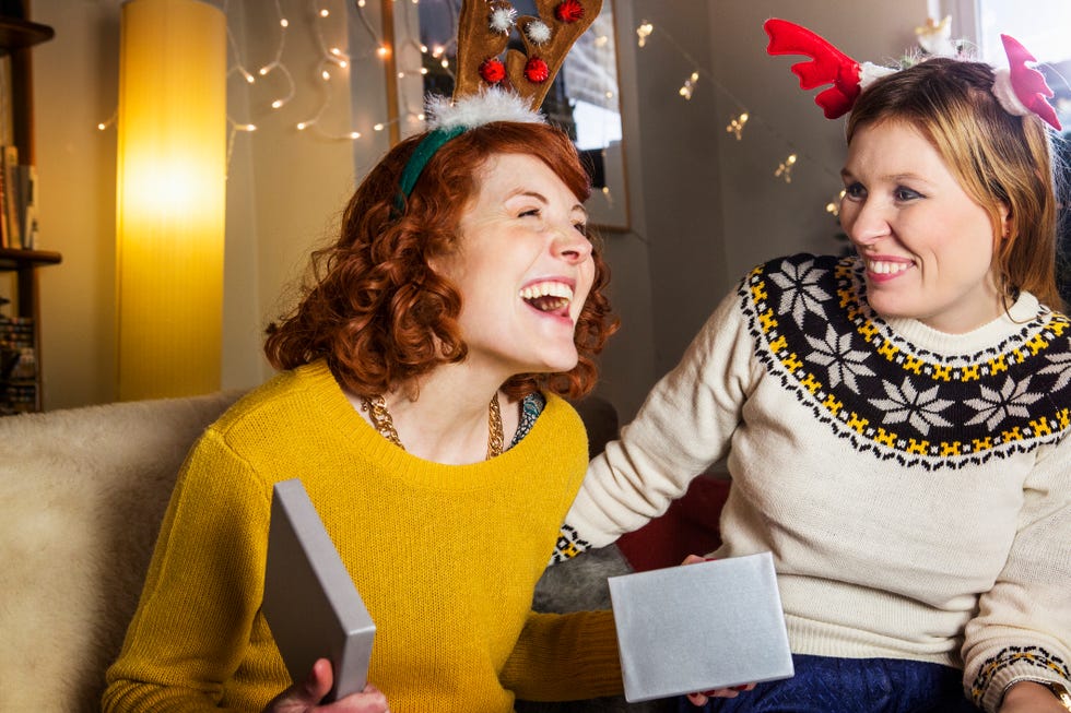 redheaded woman smiling when sitting on a couch opening a christmas present in a silver box there is a woman in a sweater beside her smiling she is wearing pink and red antlers on top of her head