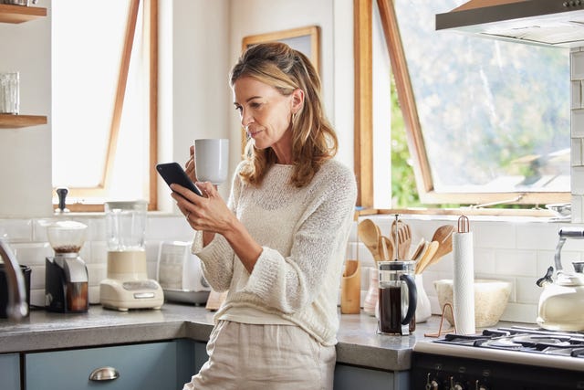 woman looking at phone while drinking a coffee in her kitchen