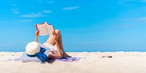 Woman Lying While Reading Book On Beach Against Blue Sky