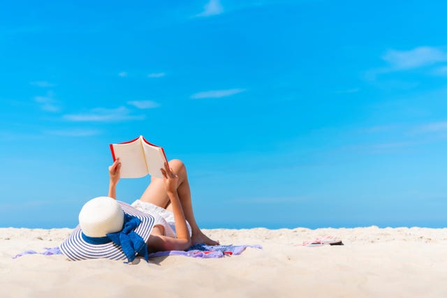 Woman Lying While Reading Book On Beach Against Blue Sky