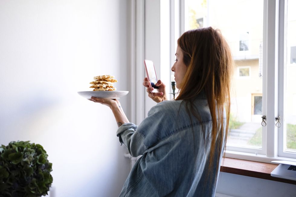 Woman photographing waffles through smart phone in plate by wall at home