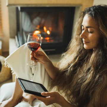 woman reading ebook on digital tablet and drinking red wine relaxing at home near fireplace