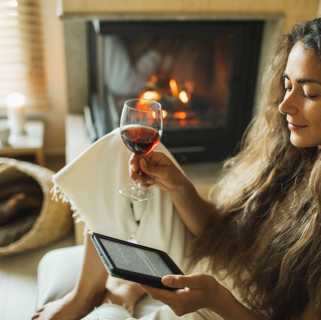 woman reading ebook on digital tablet and drinking red wine relaxing at home near fireplace