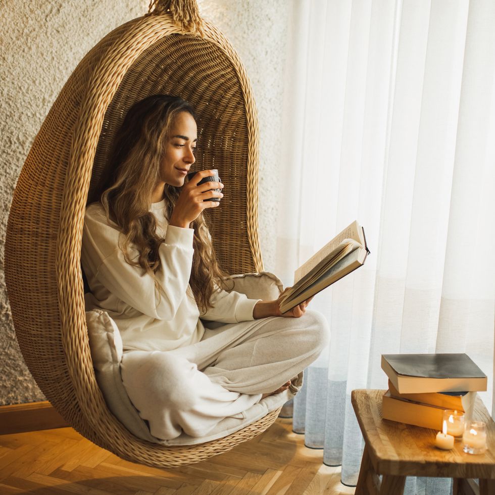 woman relaxing at home in hanging ball chair, reading book and drinking hot herbal tea