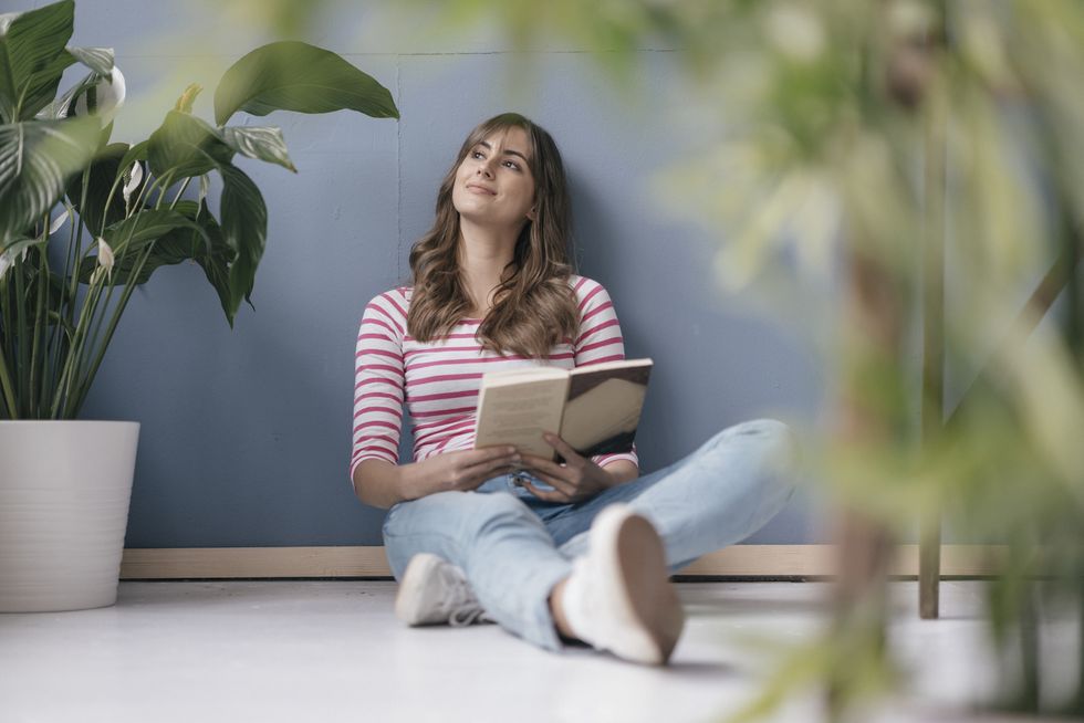 Woman sitting on ground in her new home, reading a book, surrounded by plants