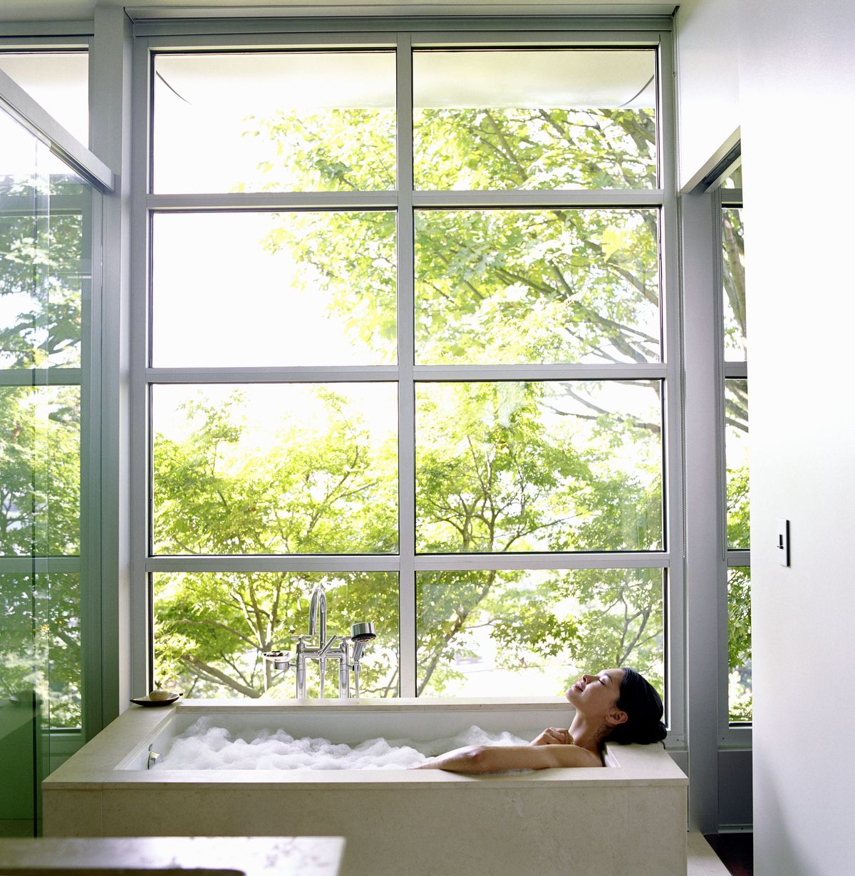 woman soaking in bubble bath, surrounded by glass windows, side view