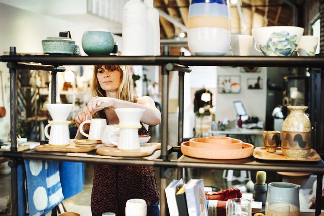 woman standing in a shop, arranging ceramic objects on a shelf.