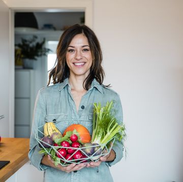 woman standing in kitchen, holding basket full of fresh fruit and vegetables