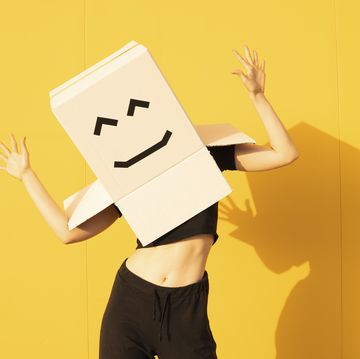 woman wearing box with smiley face gesturing in front of yellow wall