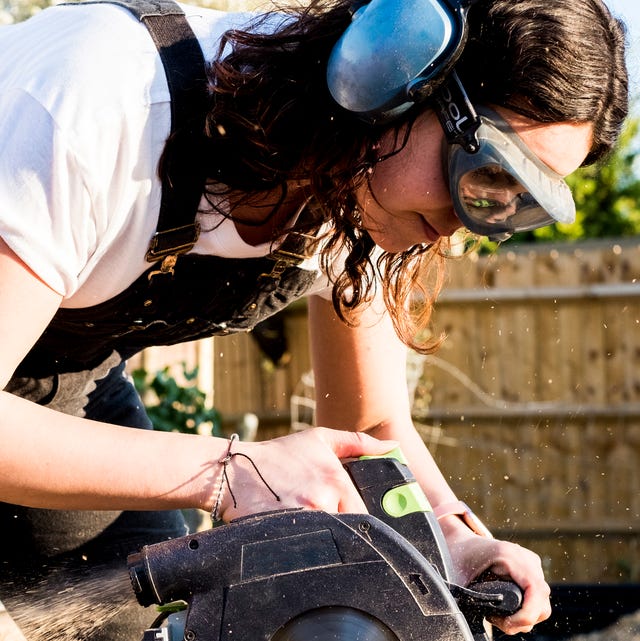 Woman wearing protective goggles and ear protectors holding circular saw, cutting piece of wood on building side.