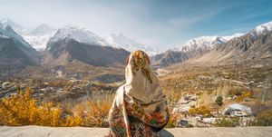 a woman wearing traditional dress sitting on wall and looking at hunza valley in autumn season, gilgit baltistan in pakistan