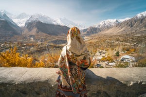 a woman wearing traditional dress sitting on wall and looking at hunza valley in autumn season, gilgit baltistan in pakistan