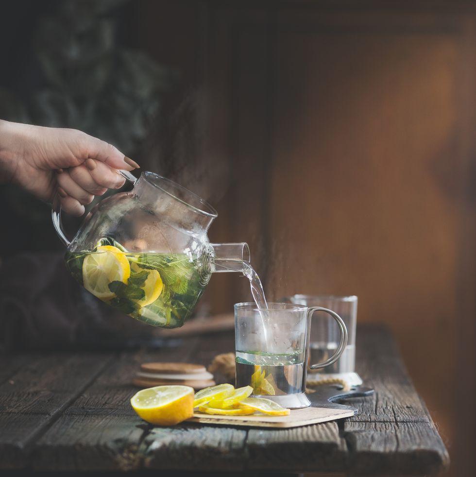 woman's hand pouring hot herbal tea from teapot in glass on rustic table with lemon slices
