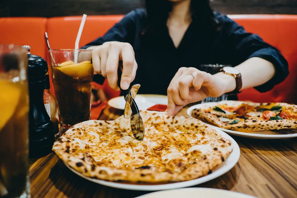 woman's hand slicing pizza with wheel cutter on table and sharing with friends in restaurant