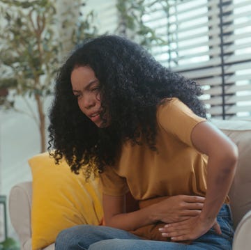 woman holds her hands to stomach in pain as she sits on sofa in office