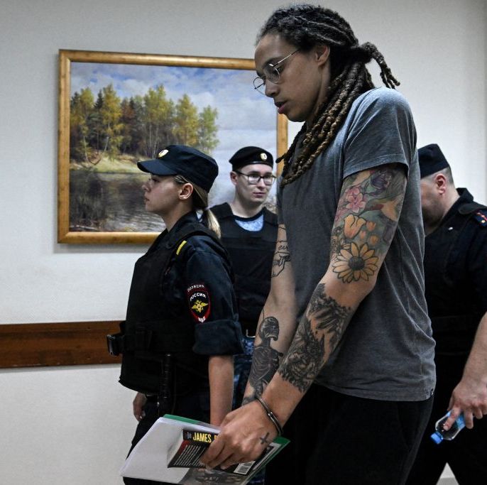 brittney griner, wearing glasses and a blue prison jumpsuit, is handcuffed and escorted down a hallway by uniformed officers
