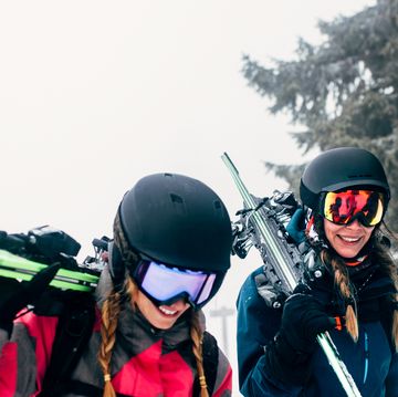 two women in jackets helmets and snow goggles carrying skis off slope