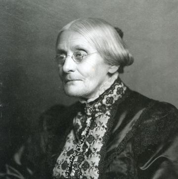 susan b anthony facing left of the camera in a black and white photo, she has a solemn expression on her face and her hair is pulled back into a low bun, she is wearing wire rimmed glasses and a fancy black satin dress with white and black lace detailing, a cameo brooch is attached to the dress collar