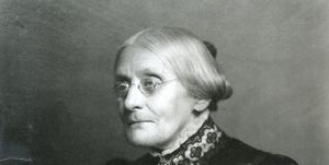 susan b anthony facing left of the camera in a black and white photo, she has a solemn expression on her face and her hair is pulled back into a low bun, she is wearing wire rimmed glasses and a fancy black satin dress with white and black lace detailing, a cameo brooch is attached to the dress collar