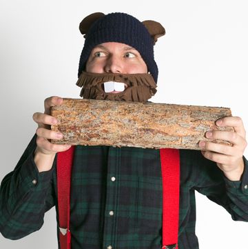 man in funny fleece woodchuck mask and ears wearing red suspenders and flannel shirt pretending to gnaw on log