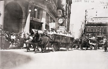 a group of women ride in a horse drawn wagon with pro union signs followed by other horse drawn buggies, a group of people watch from the sidewalk