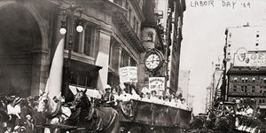 a group of women ride in a horse drawn wagon with pro union signs followed by other horse drawn buggies, a group of people watch from the sidewalk