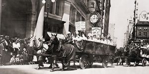 a group of women ride in a horse drawn wagon with pro union signs followed by other horse drawn buggies, a group of people watch from the sidewalk