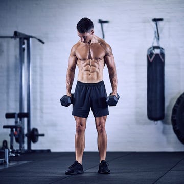 muscular male bodybuilder standing front on to camera holding dumbbells looking down in industrial gym