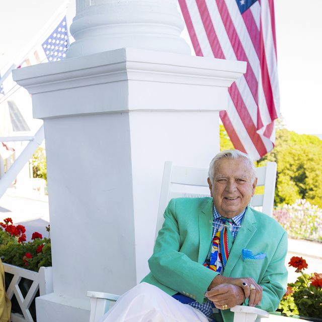 a man in a green jacket and white pants sits on a white rocking chair on a porch with an american flag in the background