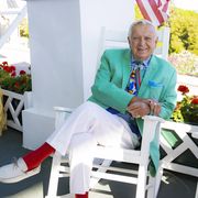 a man in a green jacket and white pants sits on a white rocking chair on a porch with an american flag in the background