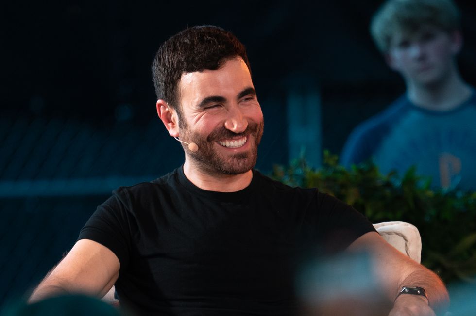 brett goldstein wears a black shirt and a white microphone headset while sitting in a chair and smiling at something off camera