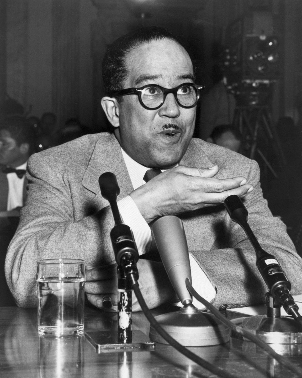 langston hughes gesturing with his right hand as he sits in front of a desk with microphones on it and speaks