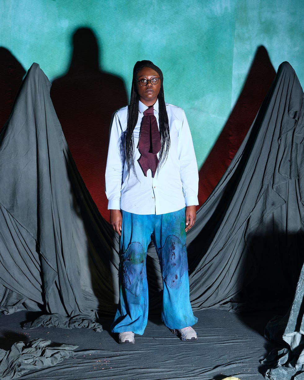 a person wearing a white shirt and blue pants with artistic designs stands in front of draped fabric